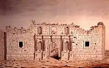 The front of a brick building; the top of the building appears torn off. Around the arched doorway are four niches, now empty. There are two small square windows.