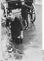 An elderly woman gathers vegetable waste tossed from a vegetable seller's wagon for her lunch, 1923