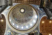 Photo looking up at the dome's interior from below. The dome is decorated at the top with a band of script. Around its base are windows through which the light streams. The decoration is divided by many vertical ribs which are ornamented with golden stars.
