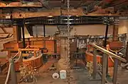 Interior view of the machinery on the first floor.  The "Great Spur Wheel" is an iron gear-wheel arranged horizontally on a vertical spindle, which spindle passes through the floor and is driven from below by the mill. on either side are millstones, set in a wooden drum-shaped housing.  They are driven by vertical iron shafts which, at the top, have a small gear mating with the great spur wheel.  The drive is thereby geared up to a much higher speed than the main vertical shaft from the wheel The floor is bare planks, the walls and apparatus are a golden coloured pale wood, and there is ancient whitewash on the walls.  The metal parts of the mechanism are painted a gloss black.