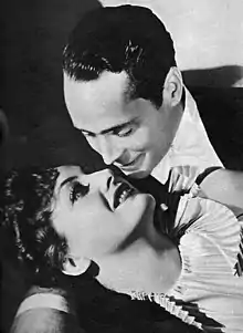 A well-dressed gentlemen embracing a coiffed woman as they look deeply into each other's eyes