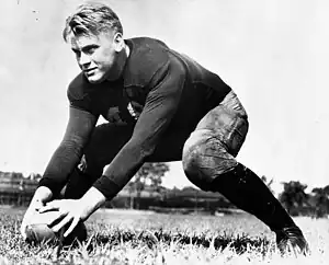 A uniformed but helmetless American Football player is shown on a football field. He is in a ready position, with legs in a wide stance and both hands on a football in front of him.