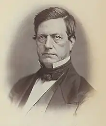 Black and white photograph of a Caucasian male with a combed-over dark hair wearing a bowtie, a black suit, and a starched white collar