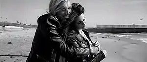 A black-and-white portrait of a brunette woman and long-haired man clinging onto each other while standing on the shore of a beach.