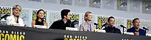 Linda Hamilton, Natalia Reyes, Diego Boneta, Mackenzie Davis, Gabriel Luna and Arnold Schwarzenegger seated at a table on a dais.