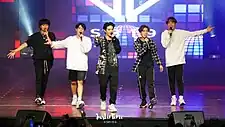 A group photo of SB19 on stage, each wearing casual clothing and holding a handheld microphone.