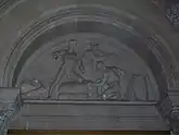 Carved stone tympanum showing a bale of cotton and two African slaves overseen by a port official leaning against a desk.