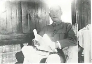 A black and white photograph of a seated man wearing a military or pseudo-military jacket