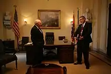  Cheney and Addington stand in front of the Wilson desk in a room with cream colored walls. A painting, and two busts hand on the wall.